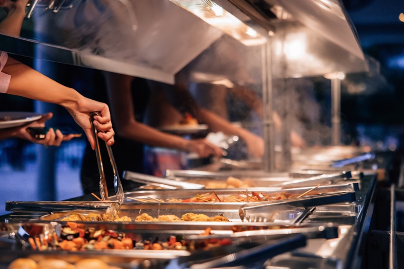 Hands of people selecting hot dishes from a steaming buffet line at an evening event, surrounded by warm lights and stainless trays risk engineering catering